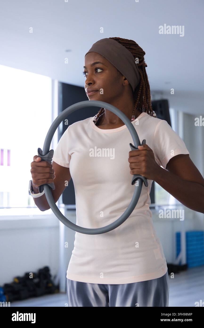 Frau hält grauen Pilatesring, während sie in einem hellen Fitnessstudio steht und ein helles Oberteil, eine Smartwatch und ein Stirnband trägt Stockfoto
