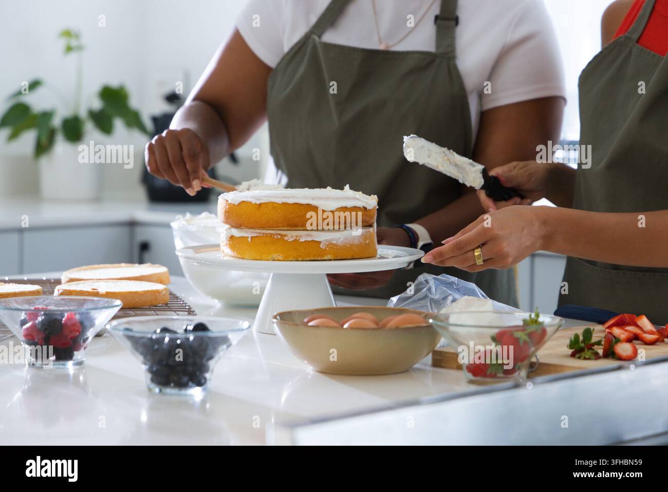 Verschiedene Freundinnen beschichten einen zweistöckigen Kuchen auf weißem Ständer an der Küchentheke mit einer Spritztüte Stockfoto