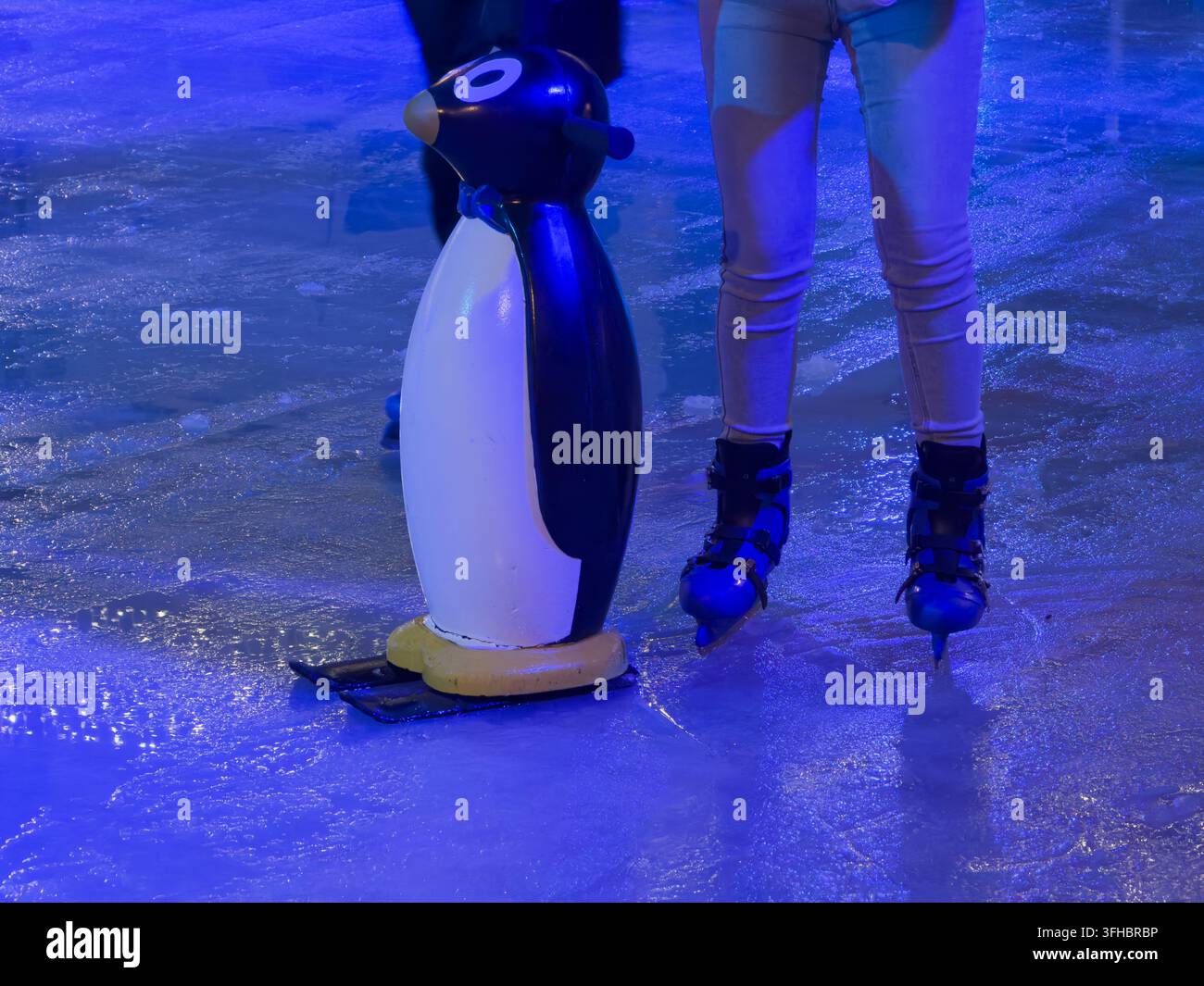 Nahaufnahme zeigt blaue Eisschlittschuhe einer Person in der Nähe einer hilfreichen pinguinformigen Stütze auf einer leuchtenden Eisfläche, die Winterfreizeit, das Erlernen des Gleichgewichts und Freude hervorruft Stockfoto