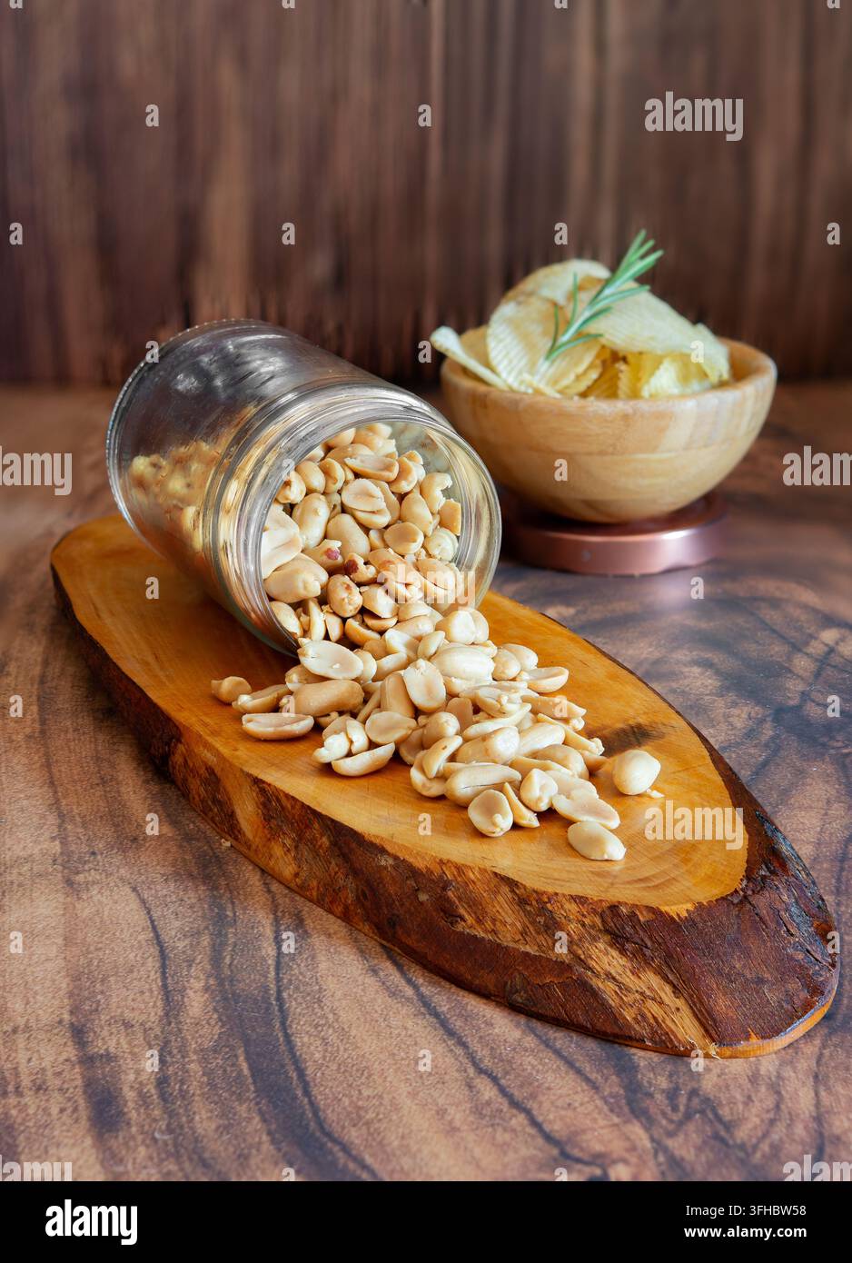 Geschälte Erdnüsse, die aus einem Gläser auf ein Holzbrett fallen, mit Kartoffelchips in einer Schüssel Stockfoto
