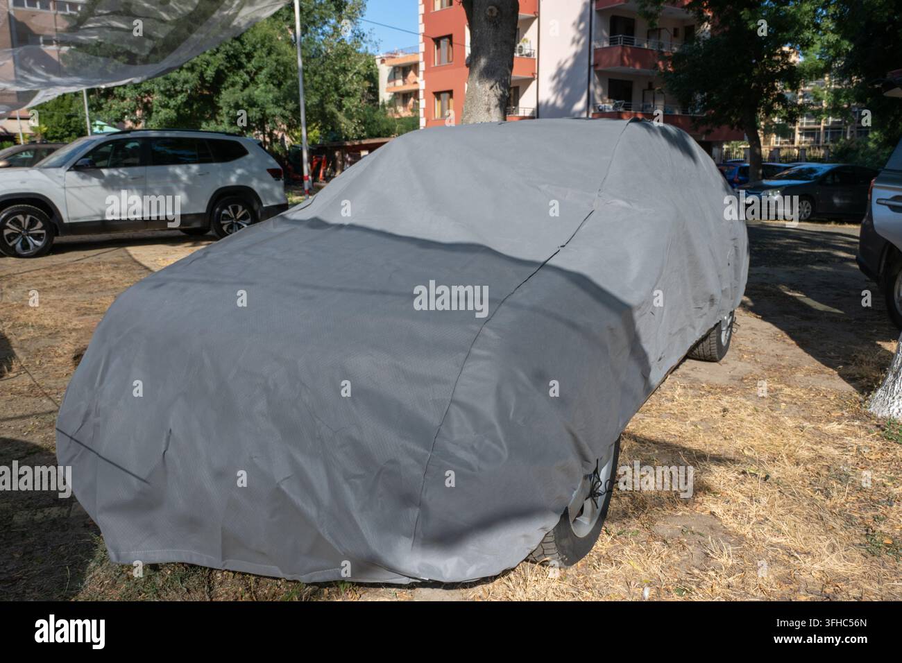 Ein geparktes Personenauto, das vollständig mit einer schützenden grauen wasserdichten Stoffplane in einem Außenparkplatz in einer Wohngegend abgedeckt ist. Stockfoto