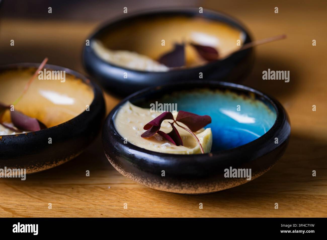 Kremige Butter mit Salzflocken überzogen und mit frischen roten Blättern garniert, serviert in handgefertigten dunklen Keramikschalen. Eine elegante Nahrungspräsentation. Stockfoto