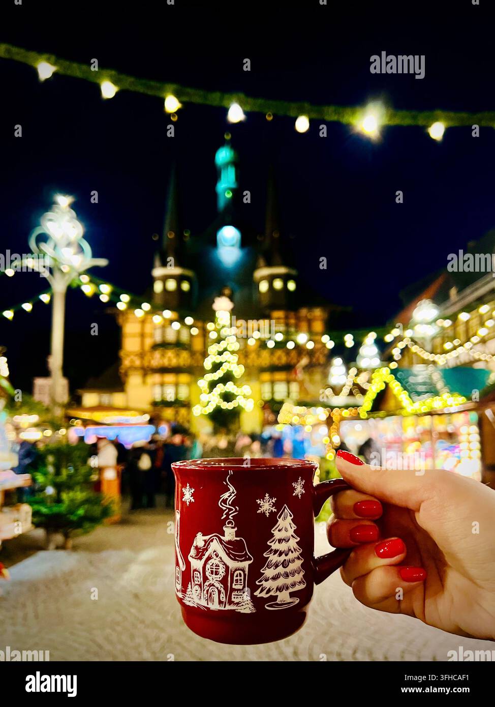 eine rote Glühweintasse auf dem Weihnachtsmarkt in Wernigerode in Deutschland, mit dem beleuchteten Rathaus sanft verschwommen im Hintergrund. - Smartphone-aufgenommenes Stockfoto
