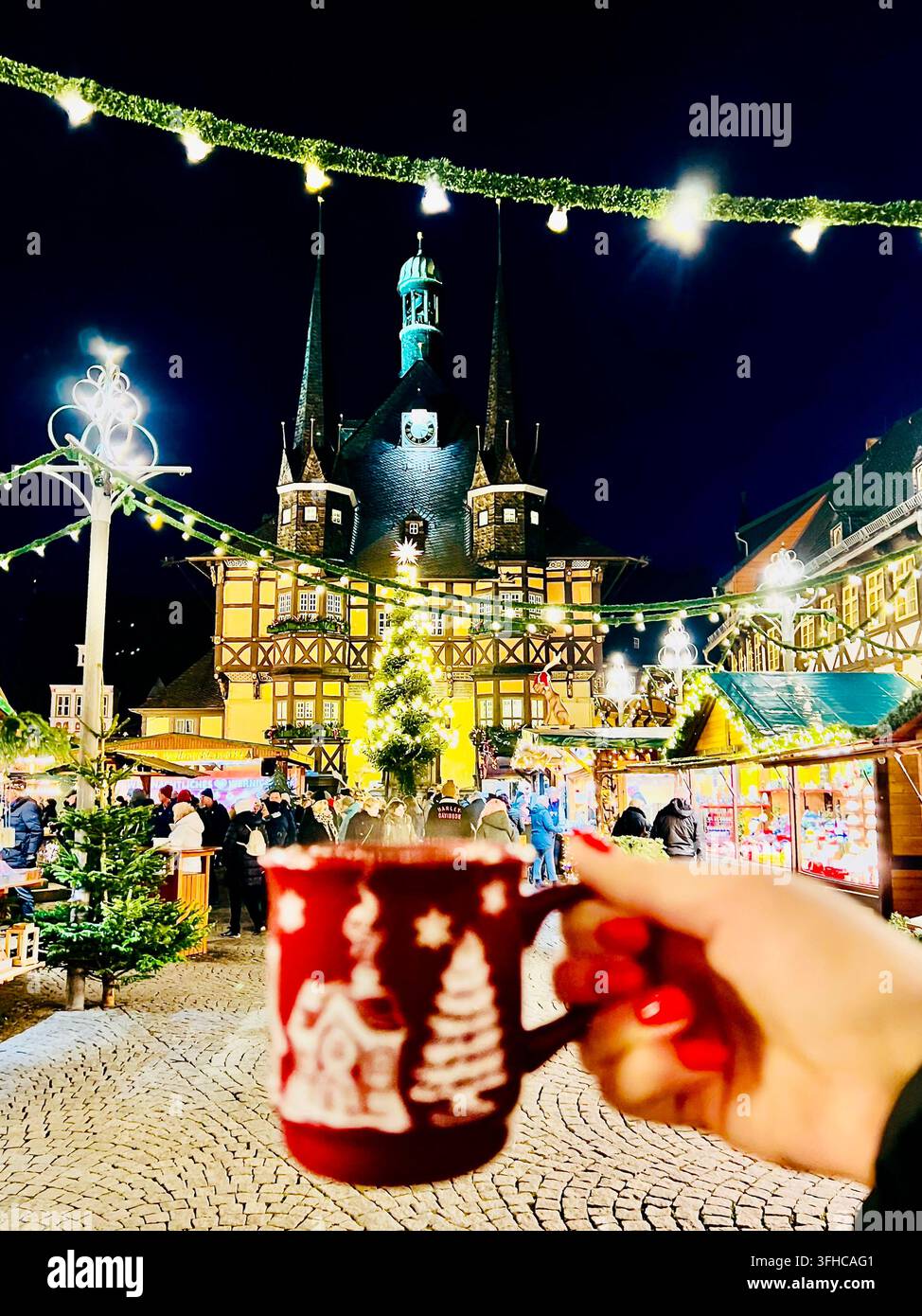 eine rote Glühweintasse auf dem Weihnachtsmarkt in Wernigerode in Deutschland, mit dem beleuchteten Rathaus sanft verschwommen im Hintergrund. - Smartphone-aufgenommenes Stockfoto