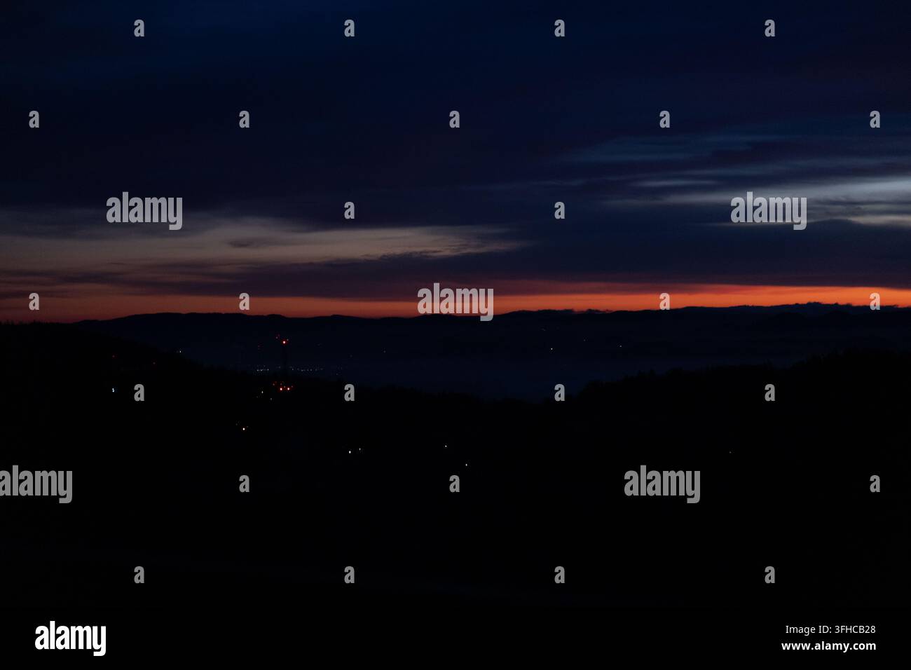 Lebendiger Abend- oder Morgenhimmel über einer dunklen Bergkette, mit leuchtenden Horizontlinien und verstreuten Lichtern abgelegener Städte, die ruhige Einsamkeit und Atmosphäre vermitteln Stockfoto