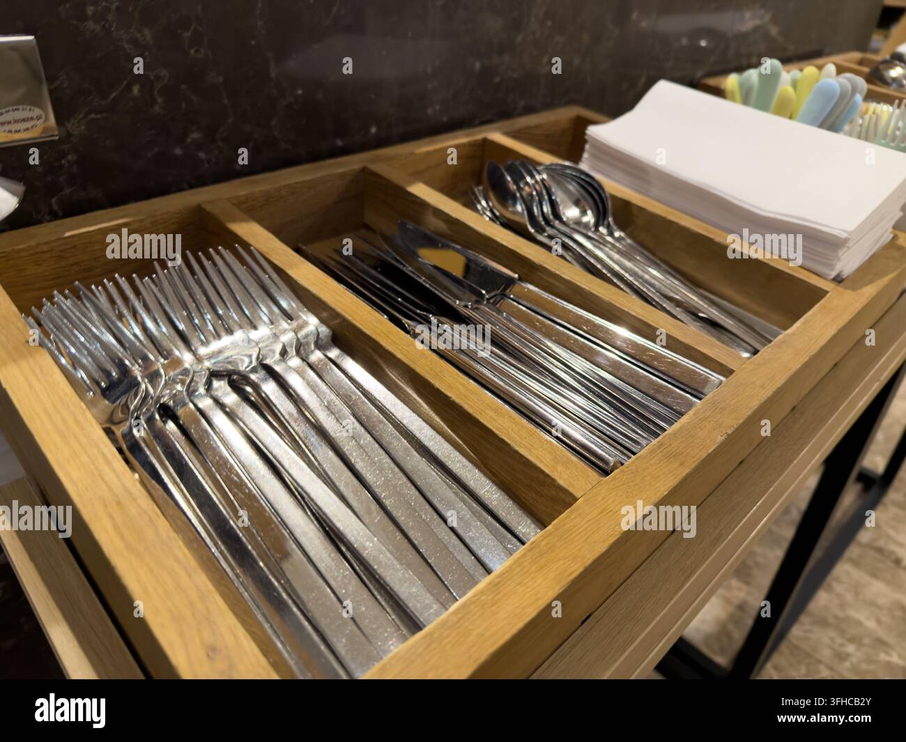 Detaillierte Aufnahme von funkelnd sauberen Edelstahlgabeln, -messern und -löffeln, die ordentlich in einem Holzhalter angeordnet sind, neben knusprigen weißen Servietten, für Hygi Stockfoto