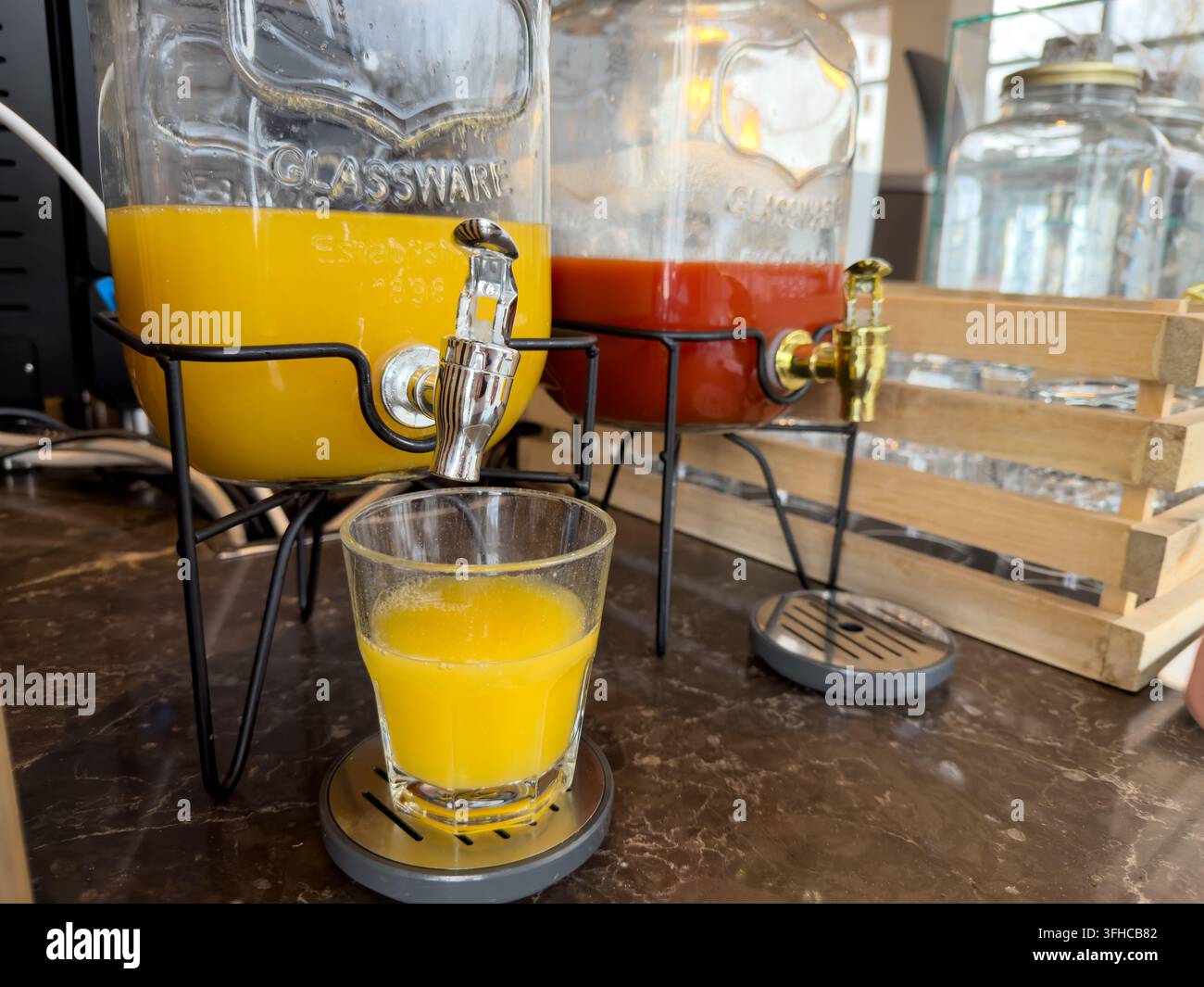 Oberansicht eines Glas-Saftspenders, der lebendigen Orangensaft in ein Glas gießt, mit einem roten Saftspender in der Nähe, ideal für Hotelfrühstück oder Catering Stockfoto