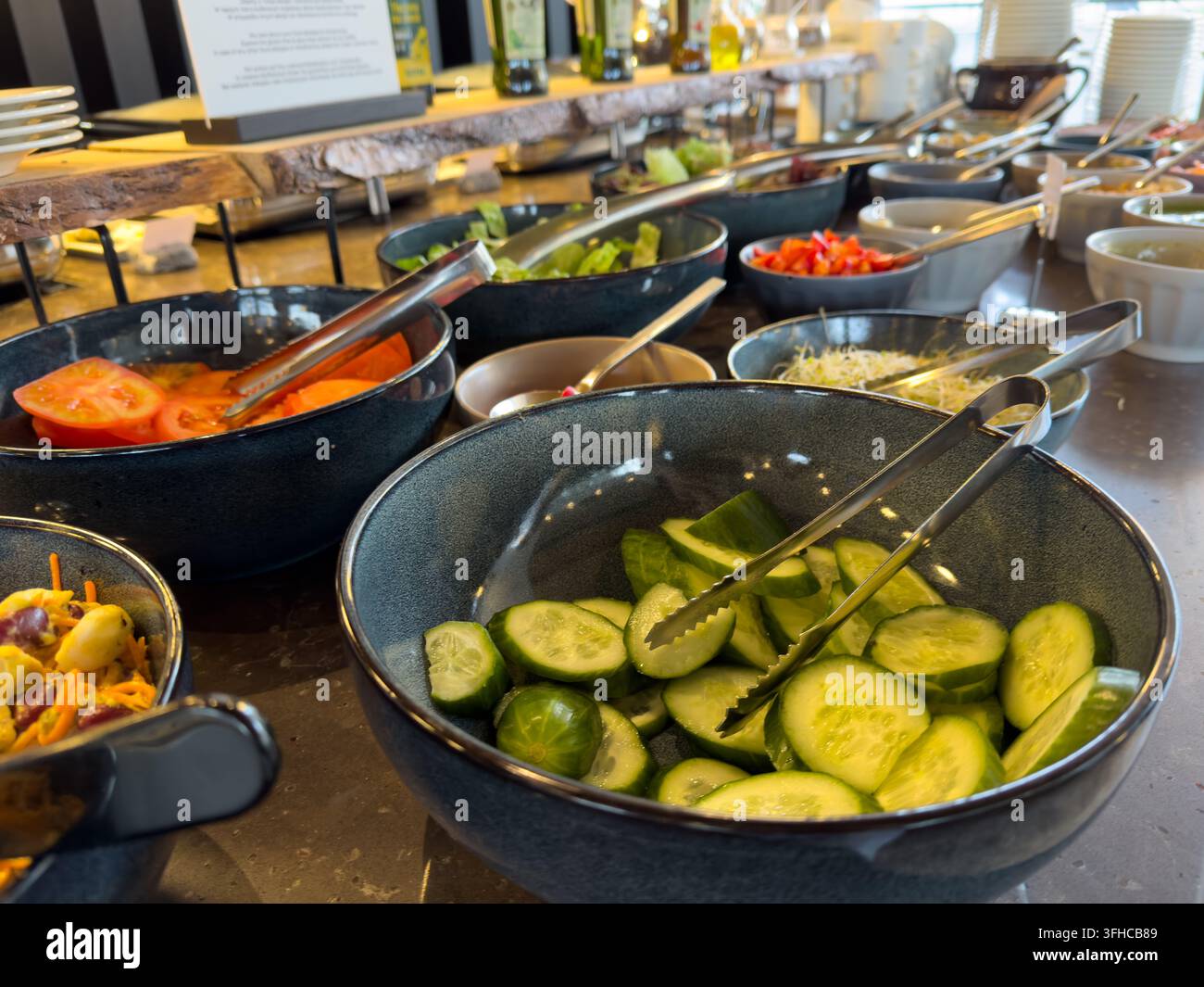 Nahaufnahme von frisch geschnittenen Gurken, Tomaten und Sprossen in Schüsseln mit Servierzangen auf einer Selbstbedienungs-Buffetlinie, die gesunde Ernährung und Catering fördert Stockfoto