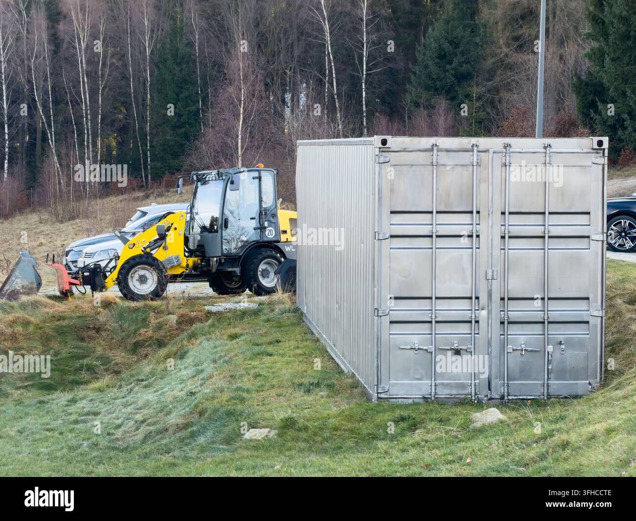 Szklarska Poręba, Polen – 16. Dezember 2025: Grauer Metall-Lagerbehälter, gelber Kompakt-Radlader: unverzichtbar für den industriellen Materialumgang, so Stockfoto