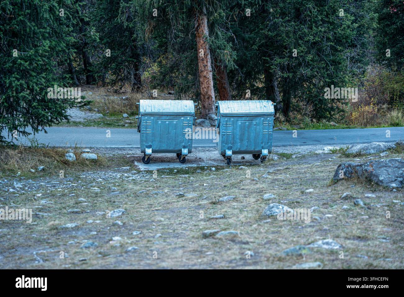 Zwei große Müllcontainer aus verzinktem Stahl mit Rädern und Deckeln stehen nebeneinander neben einer Asphaltstraße in einem bewaldeten, bergigen Gebiet. Stockfoto