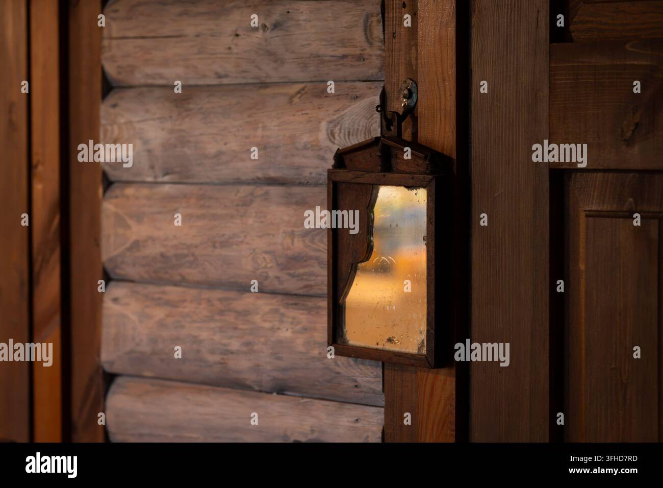Vintage-Wandspiegel, der an einer rustikalen Holzbalkenwand montiert ist, mit einem gealterten Rahmen und reflektierendem Glas. Warmes goldenes Licht, das sich im Spiegel spiegelt. Stockfoto