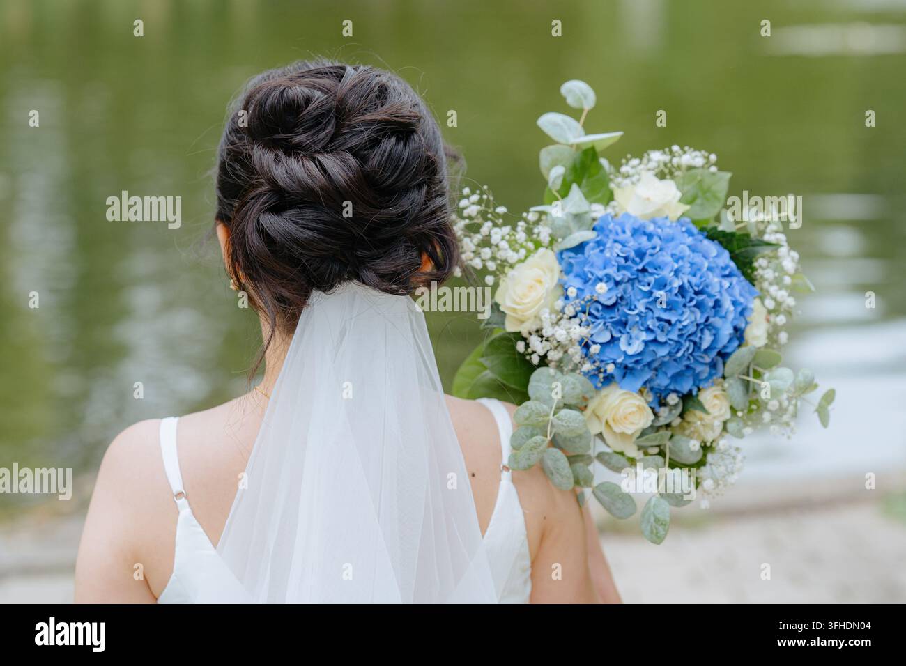 Braut hält einen Strauß, im Freien in der Nähe eines Gewässers, mit Blumenstrauß, Schleier, Hochzeitskleid, aus einer Rückansicht, mit Weiß, Blau Stockfoto