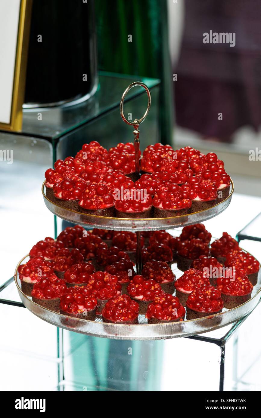 Ein mehrstufiges Desserttablett präsentiert Reihen kleiner Schokoladengehäuse, die mit glänzenden, lebhaften roten Beeren gekrönt sind. Der Ständer ruht auf einem modernen Spiegel Stockfoto