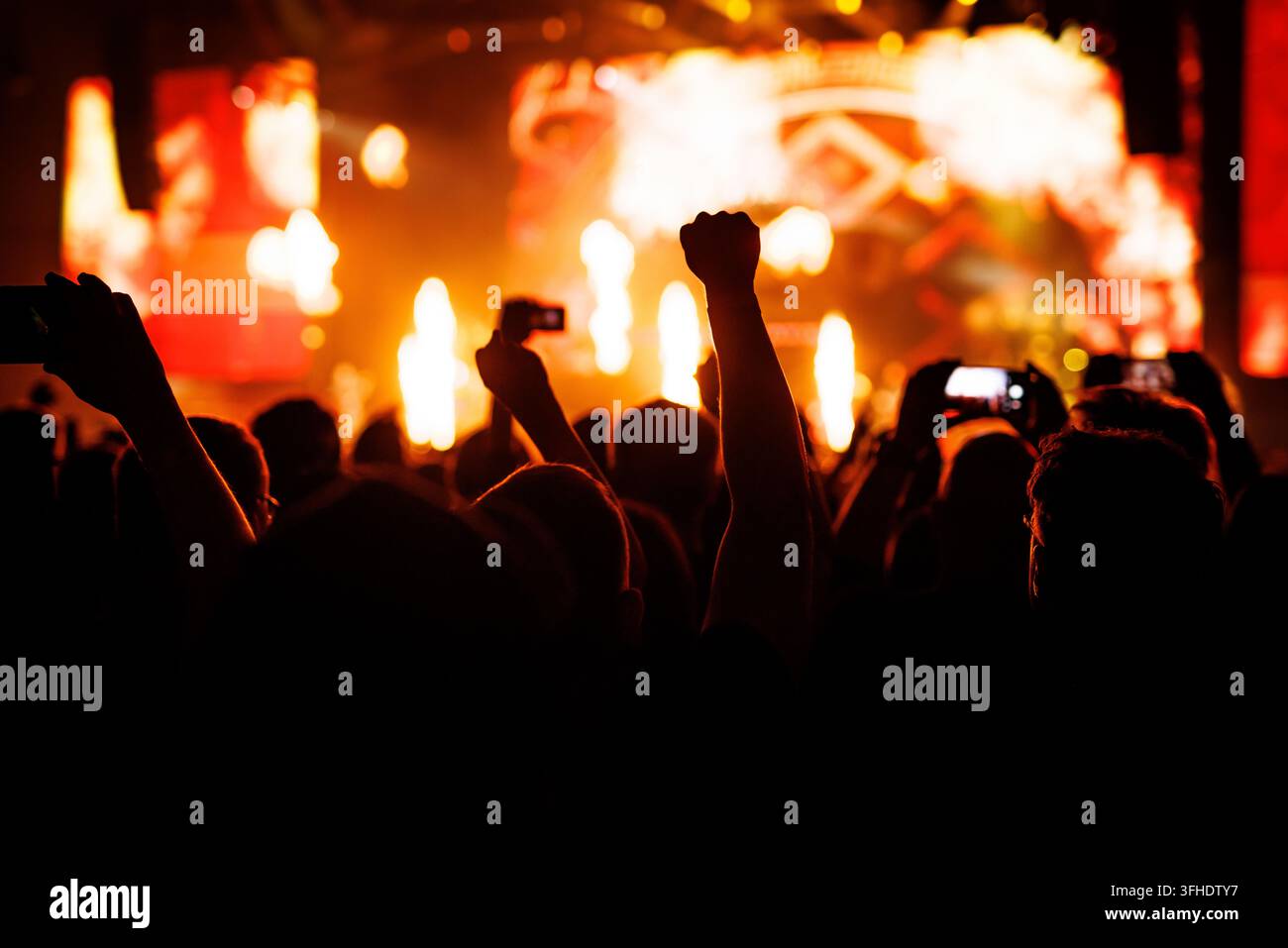 Silhouettierte Fans heben ihre Hände und Fäuste während eines energiegeladenen Rockkonzerts. Der Hintergrund zeigt explosive orangefarbene Pyrotechnik und helle Videoeffekte Stockfoto