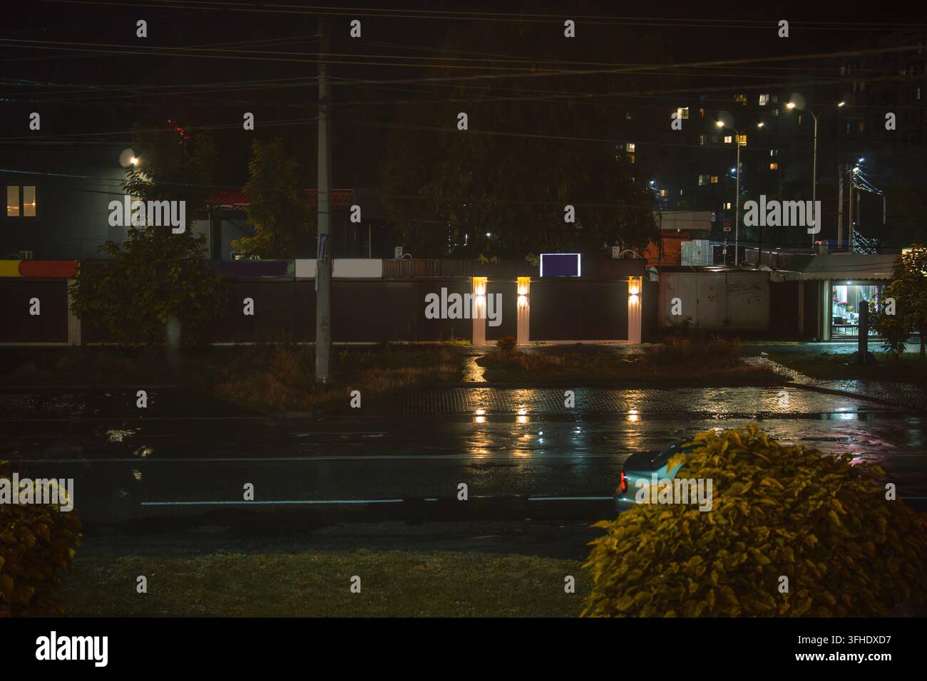 Eine horizontale Nachtaufnahme einer nassen Stadtstraße nach dem Regen. Künstliche Lichter werfen warmes Glühen und schimmernde Reflexionen auf die dunkle Asphaltstraße. Stockfoto