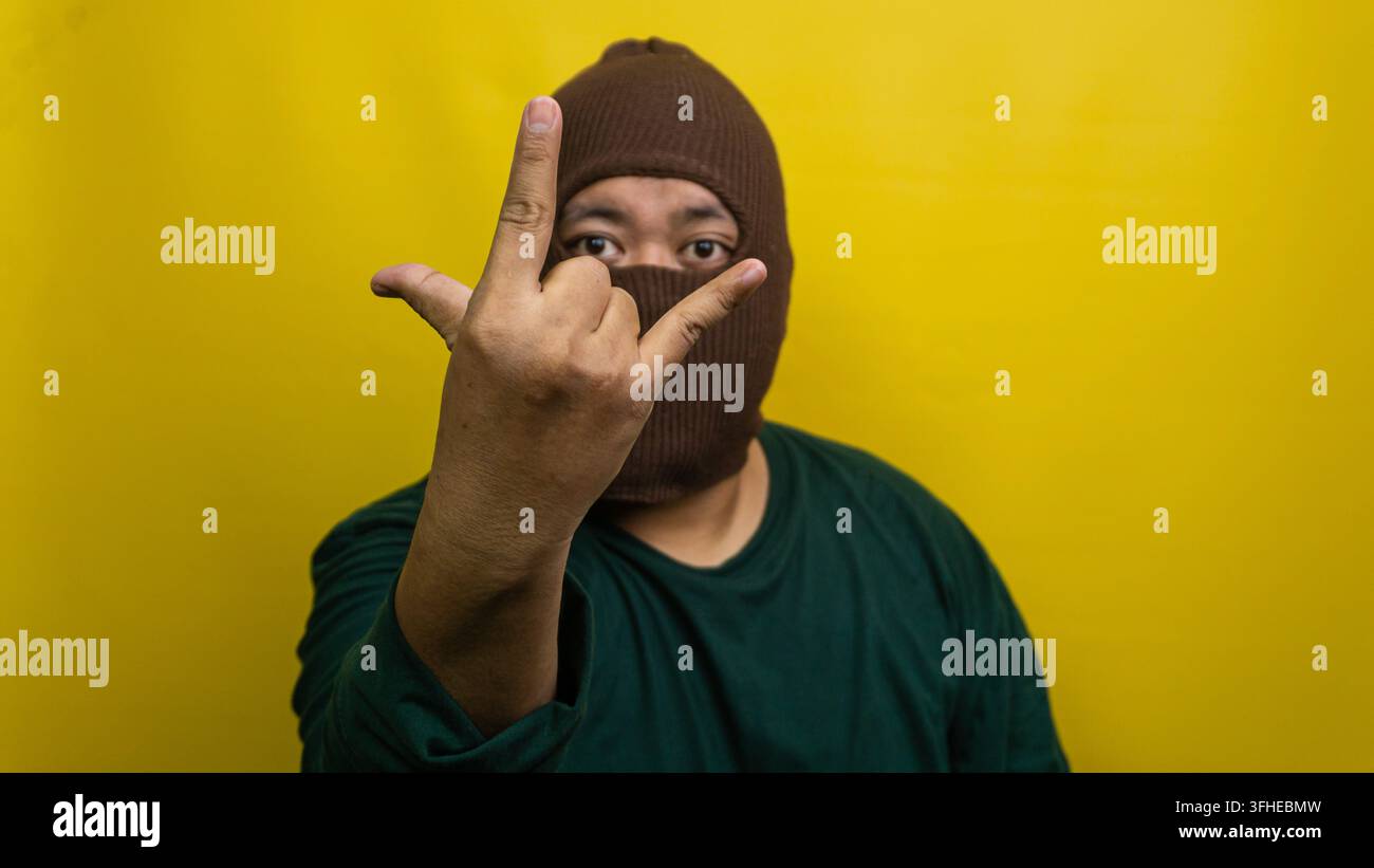 Junger asiatischer Mann mit brauner Sturmhaube zeigt Rock-and-Roll-Handgestik vor lebhaftem gelbem Hintergrund. Der maskierte Mensch streckt Zeige- und Daumen Stockfoto