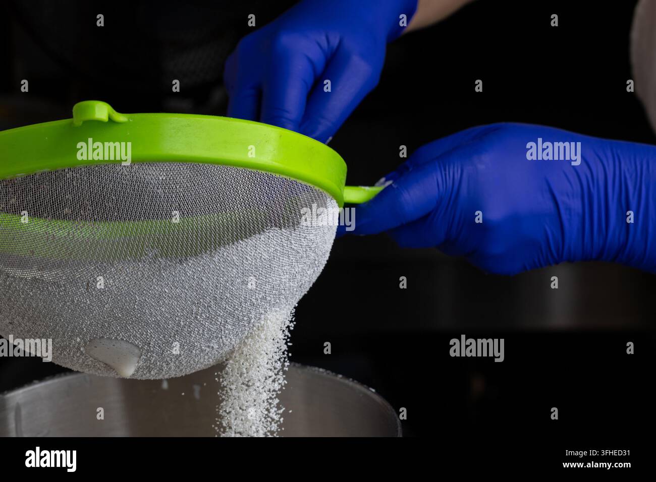 Person mit blauen Schutzhandschuhen siebt weißen granulierten Zucker durch ein feines Sieb in eine Metallschale, Nahaufnahme der Lebensmittelzubereitung Stockfoto
