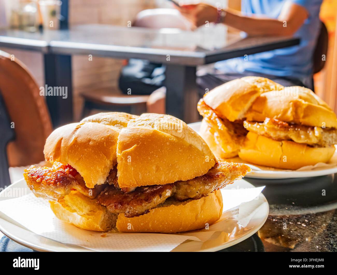 Knusprige Schweinekoteletts, die zwischen weichen Brötchen liegen, stehen auf weißen Tellern an einem geschäftigen Esstisch mit einem Kunden im Hintergrund. Stockfoto