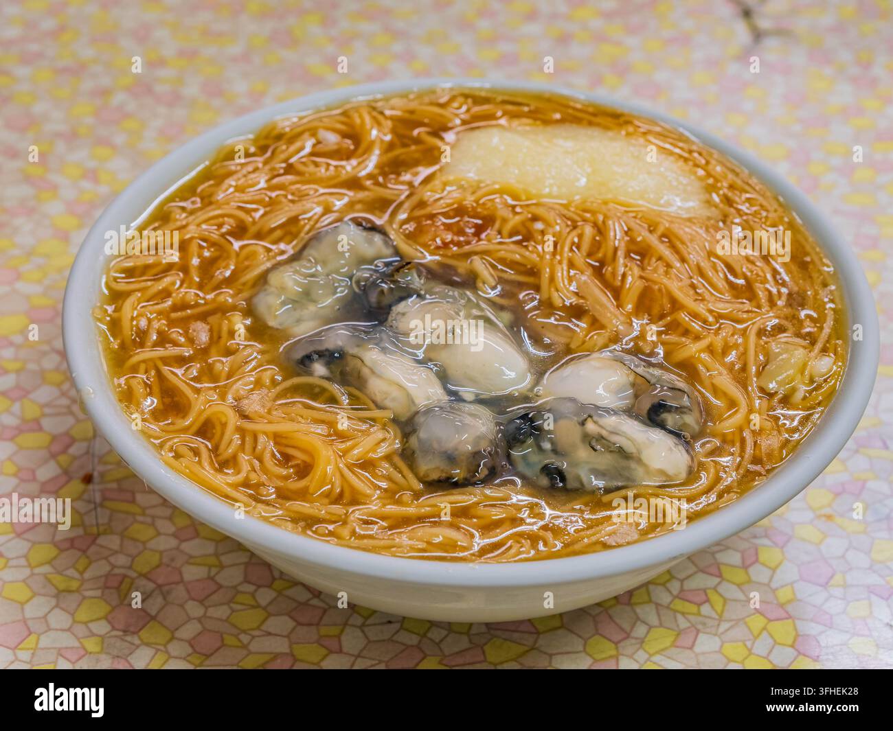 Goldene Eier-Nudeln und frische Austern ruhen in einer herzhaften Brühe in einer weißen Schüssel, die auf einer Retro-Mosaikflächchen platziert ist. Stockfoto
