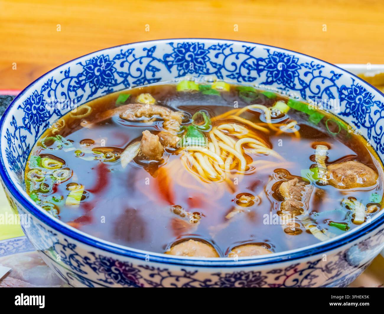 Heiße Rindfleisch-Nudelsuppe köchelt in einer blau-weiß gemusterten Schüssel mit zarten Fleischstücken, Nudeln und gehackten grünen Zwiebeln, die in einer reichhaltigen Brühe schwimmen. Stockfoto