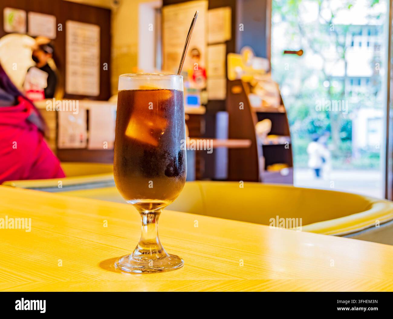Taipei, Taiwan, 14. APR 2024 – Ein hohes Glas dunkler Eistee mit einem Metallstrohhalm steht auf einer hellgelben Holzoberfläche in einem Café. Stockfoto