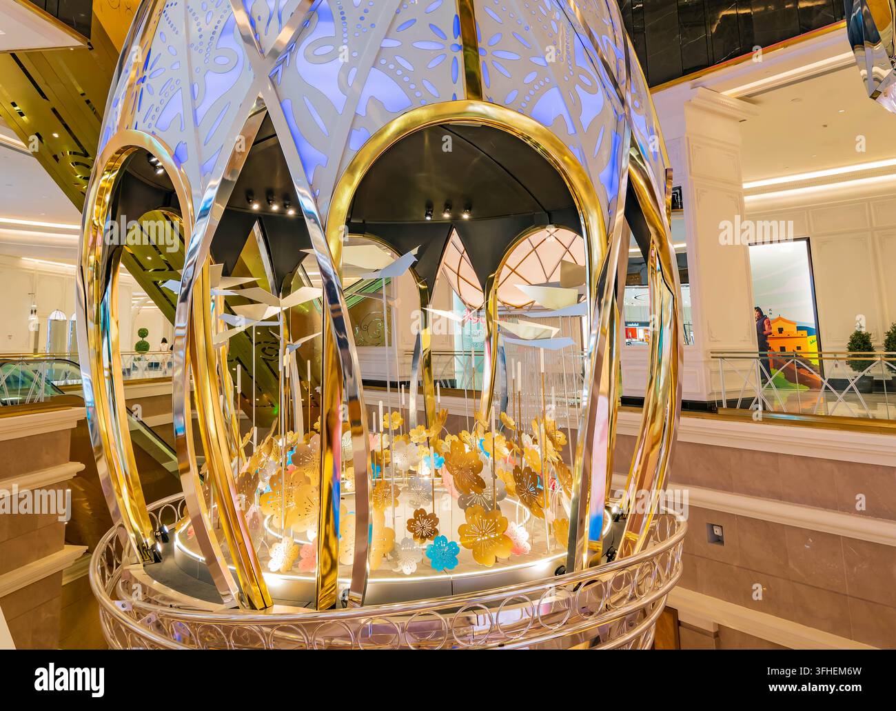 Eine kunstvolle, goldene eiförmige Installation mit blau beleuchteten Ausschnitten und hängenden Blumen steht in einem modernen Einkaufszentrums-Atrium. Stockfoto