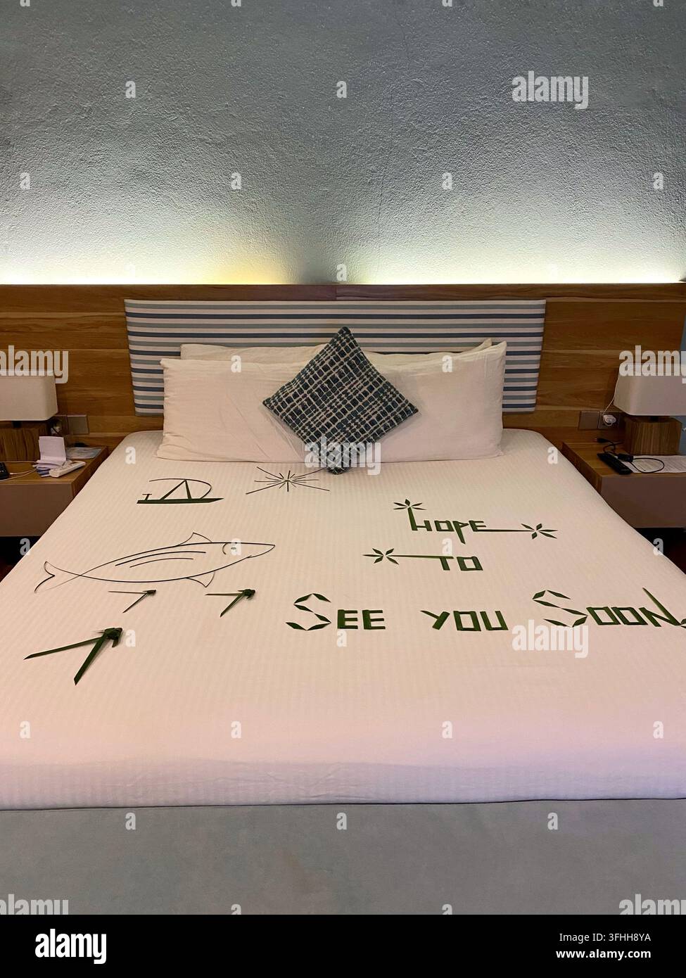 Eine Palmblatt-Abschiedsbotschaft dekoriert das Hotelbett in einem warm beleuchteten Resortzimmer. - Smartphone-aufgenommenes Stockfoto
