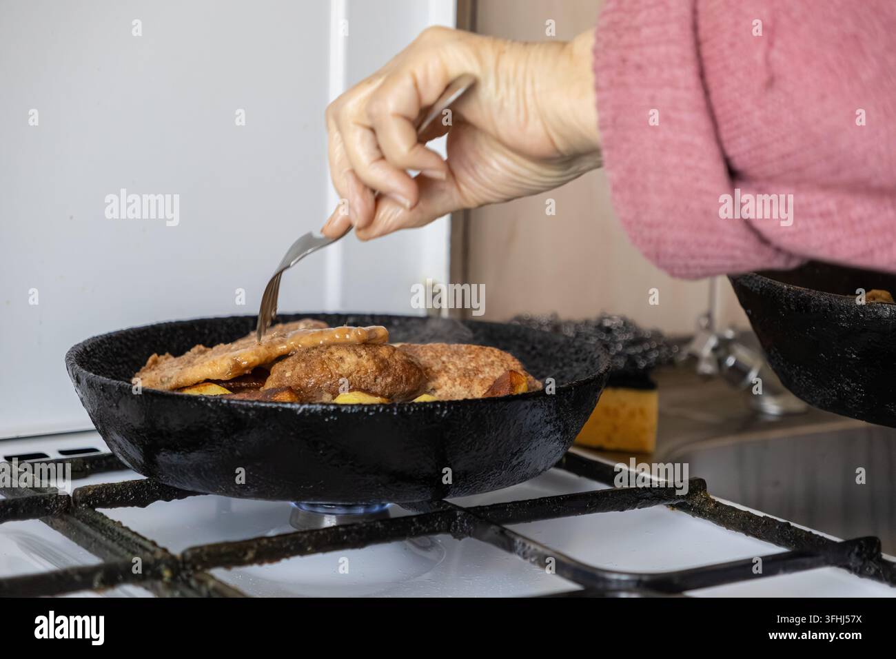Eine gealterte Hand verwendet eine Gabel, um gebräunte Schweinekoteletts in einer schweren Gusseisenpfanne zu wenden, was ein Gefühl von Hausmannskost, authentischen kulinarischen Traditionen und einem Stockfoto