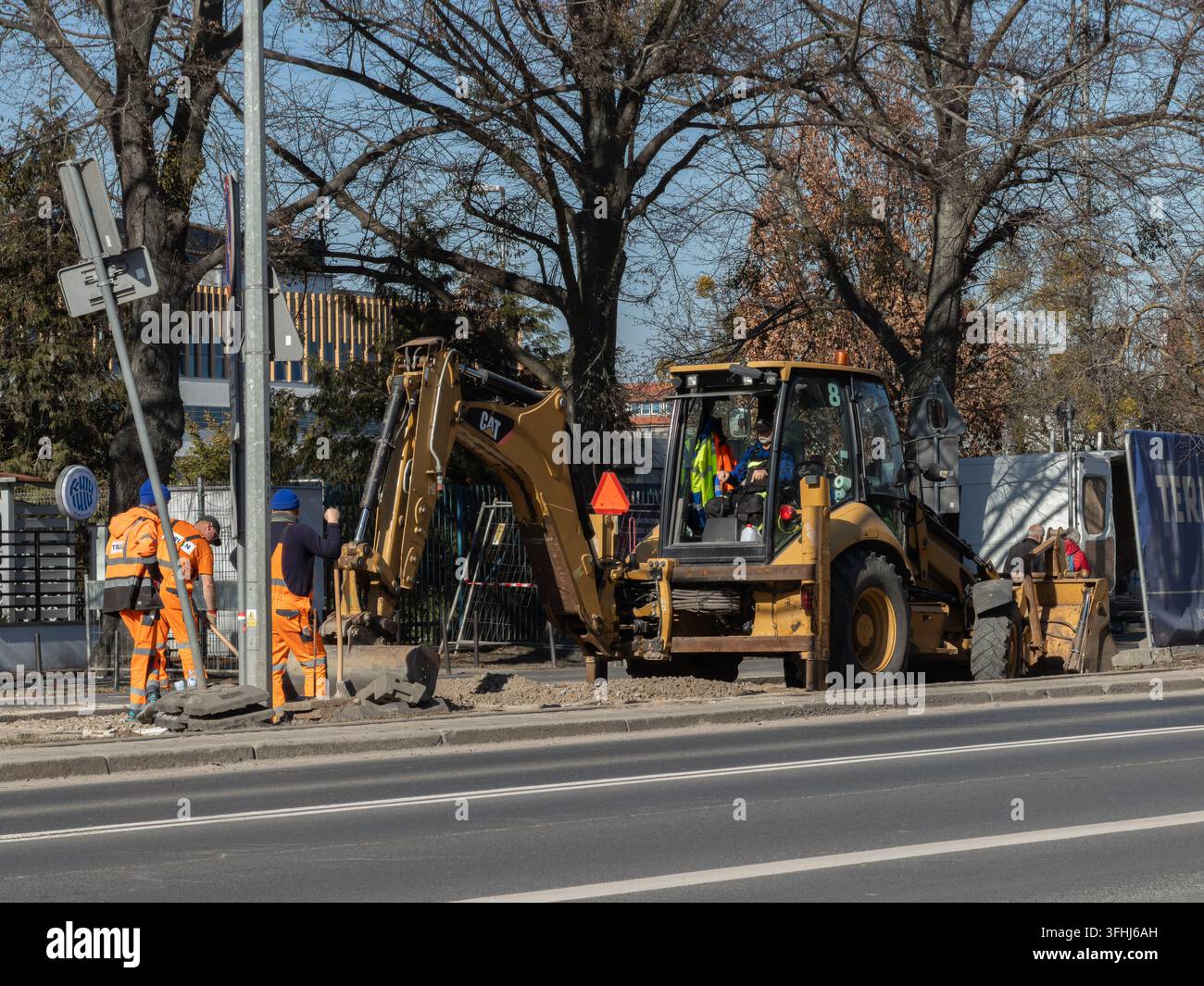 Posen, Polen – 6. März 2026: Caterpillar-Baggerlader, der neben Arbeitern in reflektierenden orangefarbenen Uniformen geparkt ist, die das Straßenpflaster reparieren, um die ur Stockfoto