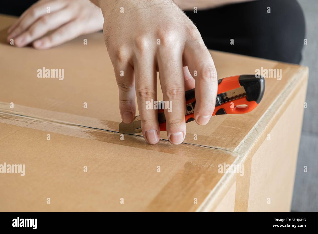 Eine Person schneidet mit einem Kartonschneider durch Paketband, um einen großen Wellpapierbehälter zu öffnen, und veranschaulicht den Prozess des Warenempfangs oder Stockfoto