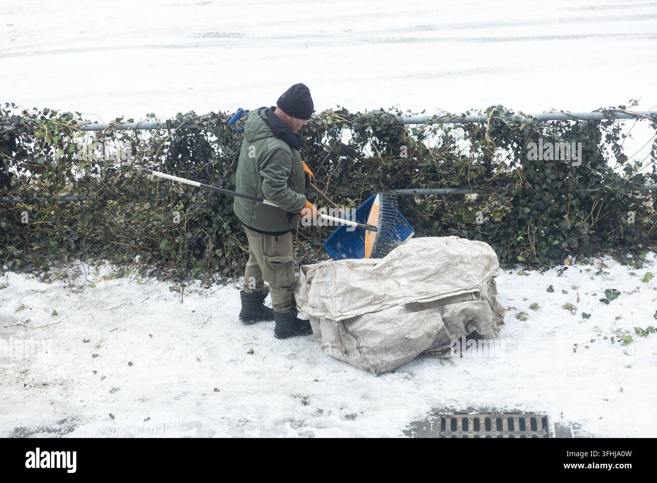 Arbeiter räumt einen verschneiten Gehweg mit einer Plastikschaufel und lädt das Eis in einen schweren Sack, was die harte Winterwartung im Freien hervorhebt, während ein f Stockfoto
