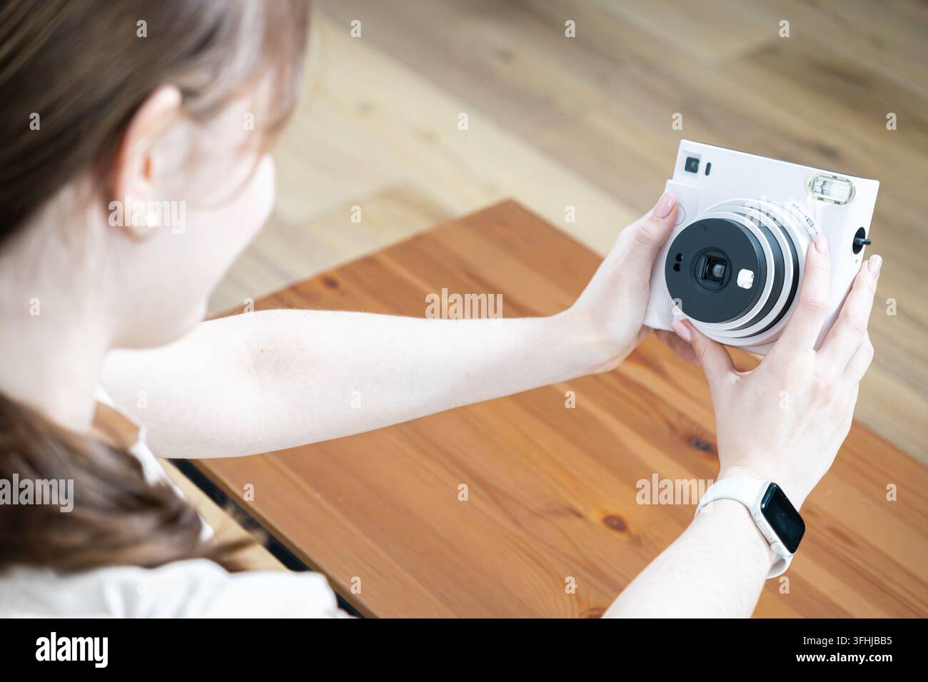 Eine Person zielt mit einer modernen Sofortbildkamera, während sie an einem Holztisch sitzt, und betont eine lässige Fotosession, die die manuelle kreative Ausdrucksweise feiert Stockfoto