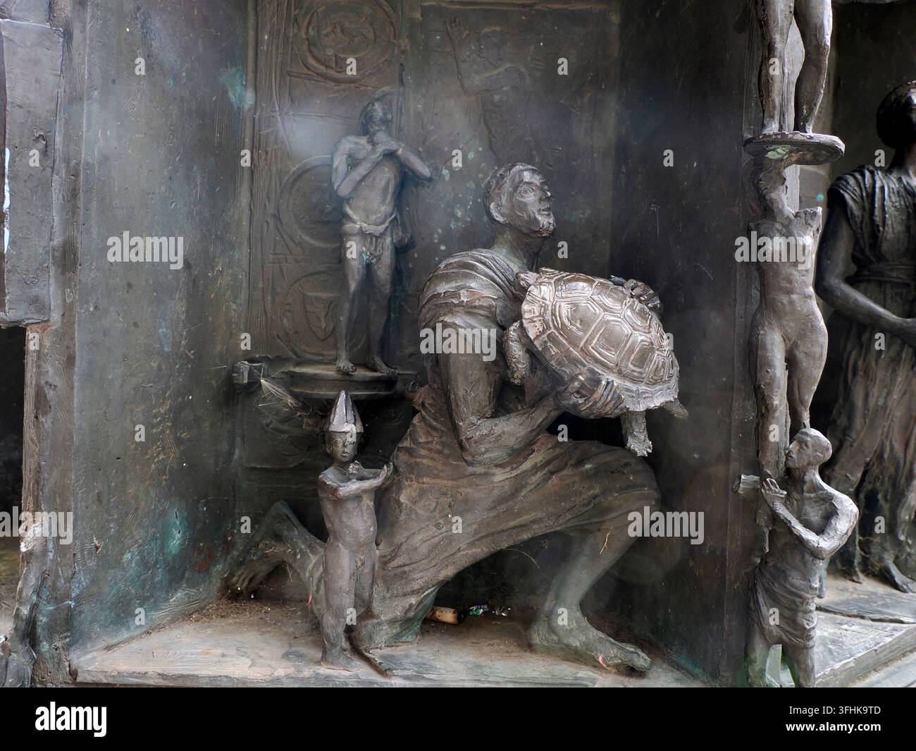 Detaillierte bronzene Skulpturenkomposition mit einem knienden Mann, der eine Schildkröte hält, kleinen Figuren und Reliefplatten in Marburg, Deutschland. Stockfoto