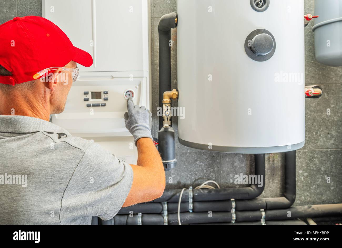 Ein Techniker zeigt auf den Druckmesser an einem wandmontierten Kessel neben einem Speicherzylinder und isolierten Rohrleitungen. Stockfoto