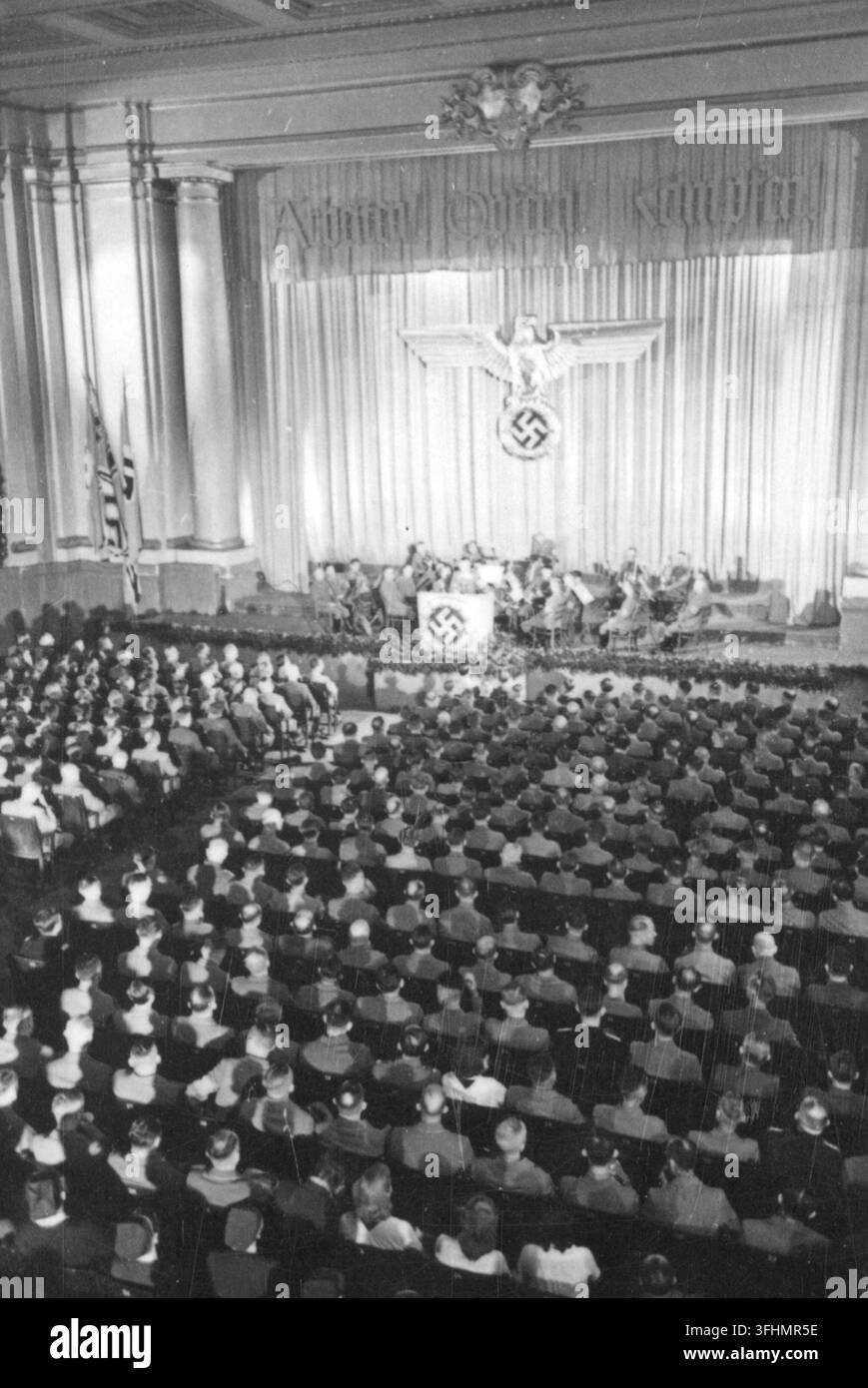 Eine große Innenversammlungshalle im Jahr 1943, mit einer Gruppe von Personen auf einer erhöhten Bühne hinter einem langen Tisch und einem Publikum, das meist dunkle Uniformen mit Mützen trägt, das der Bühne zugewandt ist, wo ein Vorhang einen blassen Vogel mit ausgebreiteten Flügeln über einem dunklen eckigen Symbol auf einem blassen Diskus zeigt, mit dem Text „Aujah! Quill! Kultur!“ darüber sichtbar. Stockfoto
