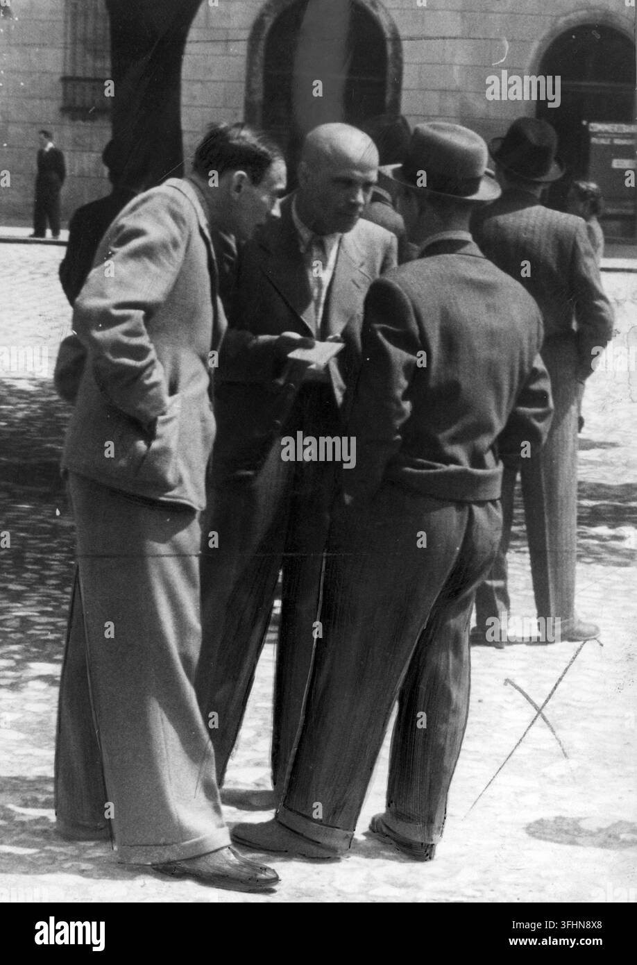 Drei arbeitslose Männer stehen zusammen auf einer Straße, einer hält ein gefaltetes Papier, vor einem großen Steingebäude mit gewölbten Türen, Juni 1941. Stockfoto
