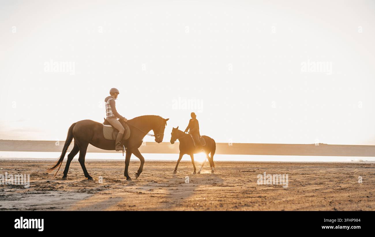 Blick auf Frauen, die Pferde entlang des Flusses in goldenem Licht bei Sonnenuntergang oder Sonnenaufgang reiten. Hengst beim Wandern in der Wüste am Wasser. Stockfoto