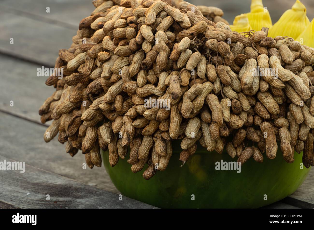 Nahaufnahme eines Haufens ungeschälter roher oder gekochter Erdnüsse und Mais in einer grünen Schüssel, die auf einem rustikalen Holztisch platziert ist. Stockfoto