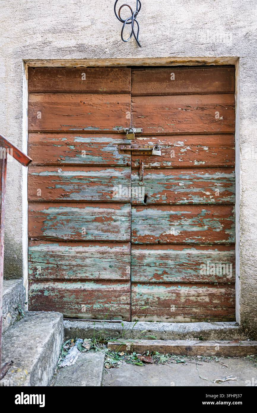 Ein altes Holztor mit abblätternder türkiser und brauner Farbe, gesichert durch rostige Vorhängeschlösser und Riegel, eingebettet in einen verwitterten Stuckrahmen mit Beton Stockfoto