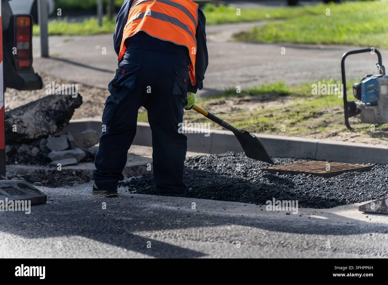 Ein Bauarbeiter bearbeitet frischen Asphalt neben einem Bordstein und einem Regenwasserabfluss, was das Ausbessern von Straßen und die Wiederherstellung der Oberfläche um Entwässerungen herum darstellt Stockfoto