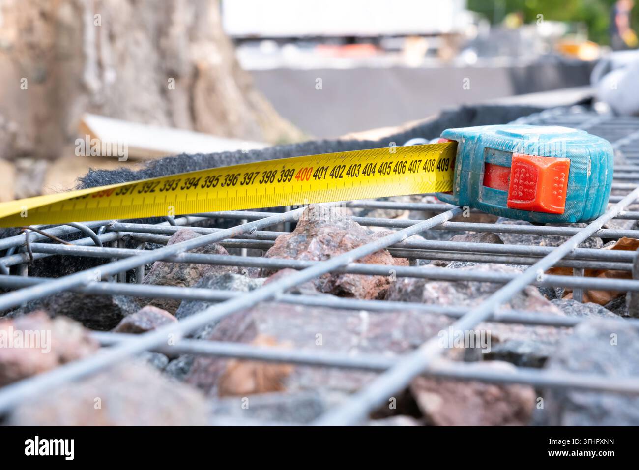 Eine Nahaufnahme eines offenen gelben Maßbands, das auf einer Stahlgitter-Gabionwand liegt, die mit Granitsteinen gefüllt ist, an einer Baustelle im Freien Stockfoto