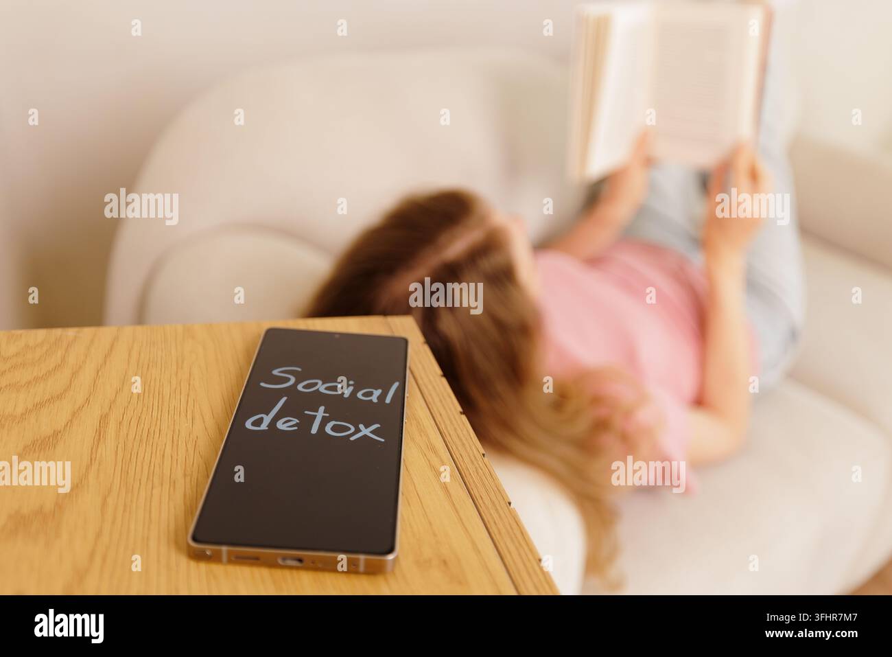 Smartphone zeigt eine Social-Detox-Nachricht auf einem Tisch, während sich eine junge Frau auf einem Sofa entspannt und ein Buch liest, was digitale Entgiftung und das Abkoppeln veranschaulicht Stockfoto