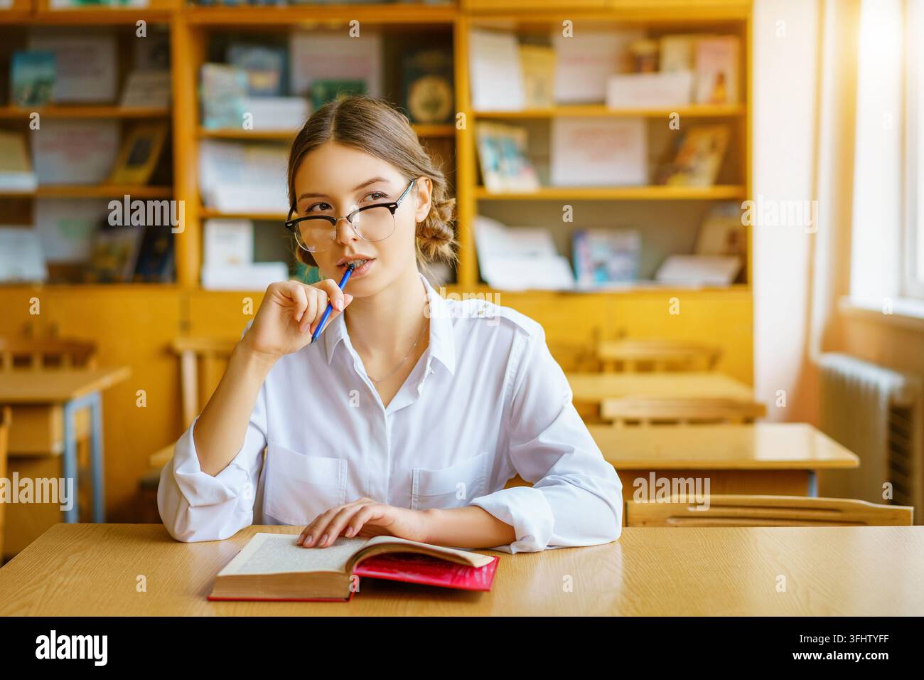 Mädchen mit Brille an einem Tisch sitzen mit einem Buch im Klassenzimmer, mit einem Bleistift in der Hand, durchdachte Suchen Stockfoto
