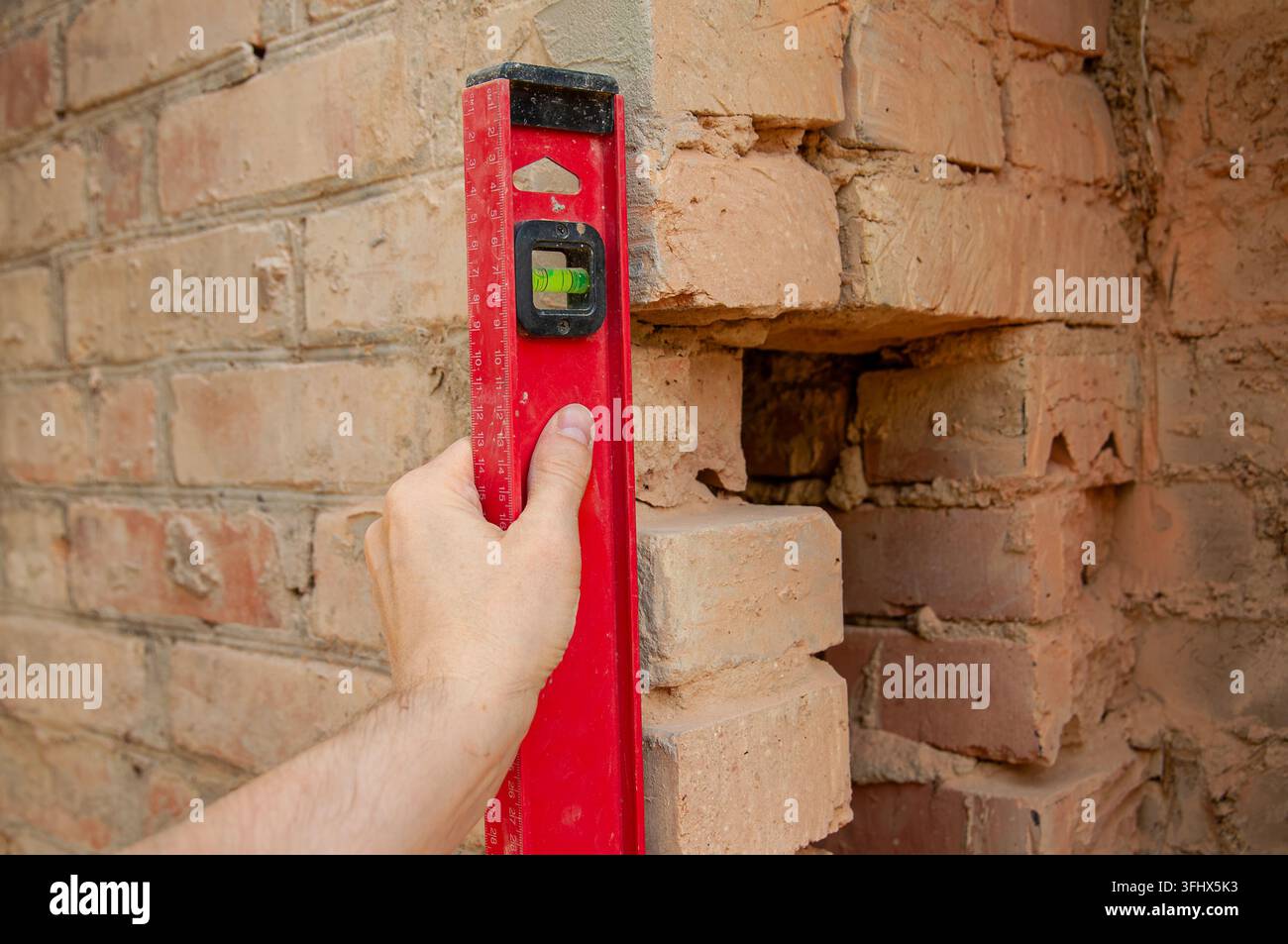 Hand hält ein Wasserwaage an einer beschädigten Ziegelmauer während Reparaturarbeiten Stockfoto