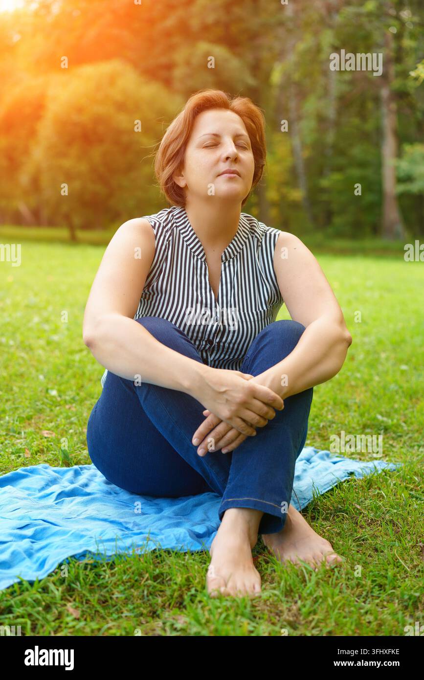 Eine erwachsene Frau sitzt auf einer Matte auf dem grünen Gras in einem Park an einem sonnigen Sommertag Stockfoto