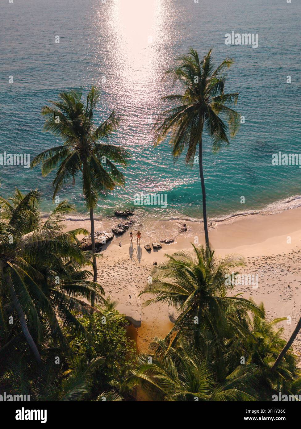 Goldenes Sonnenlicht glitzert auf den sanften Wellen, während ein Paar Hand in Hand entlang des unberührten Sandes von Takhian Beach geht, umgeben von üppigen Palmen. Stockfoto