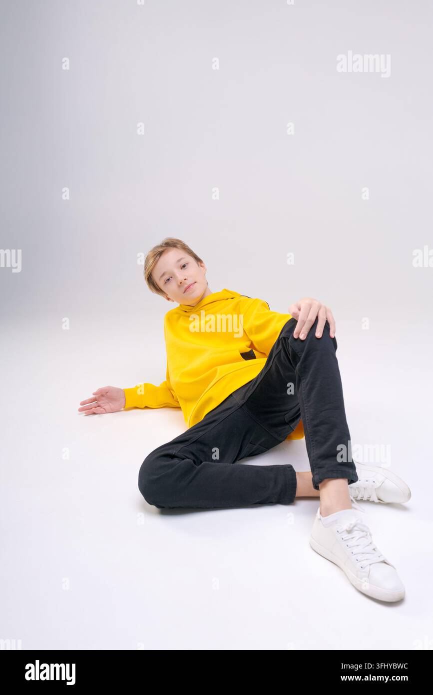 Junger, gutaussehender, glücklicher, lächelnder Teenager in schwarzen Jeans, hellgelbem Sweatshirt mit Kapuze, freut sich, während er auf einem hellen Hintergrund sitzt und posiert. Th Stockfoto