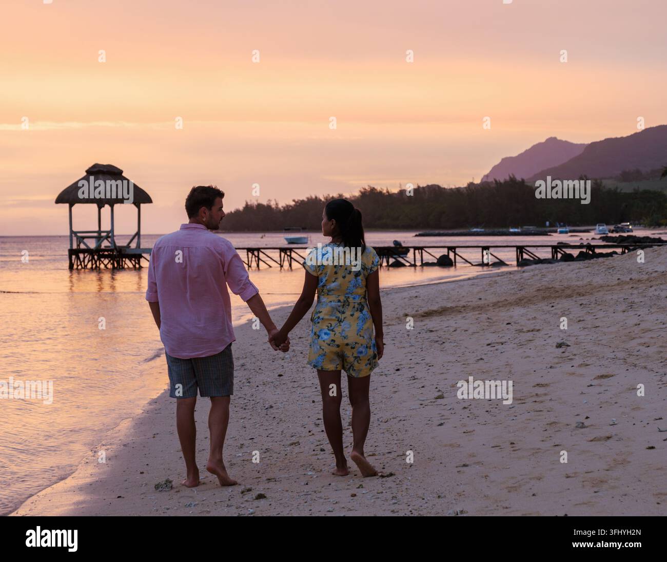 Ein paar Spaziergänge Hand in Hand an einem ruhigen Strand in Mauritius, umgeben von atemberaubenden Sonnenuntergangsfarben. Die Atmosphäre strahlt Romantik aus, perfekt für einen Luxus Stockfoto