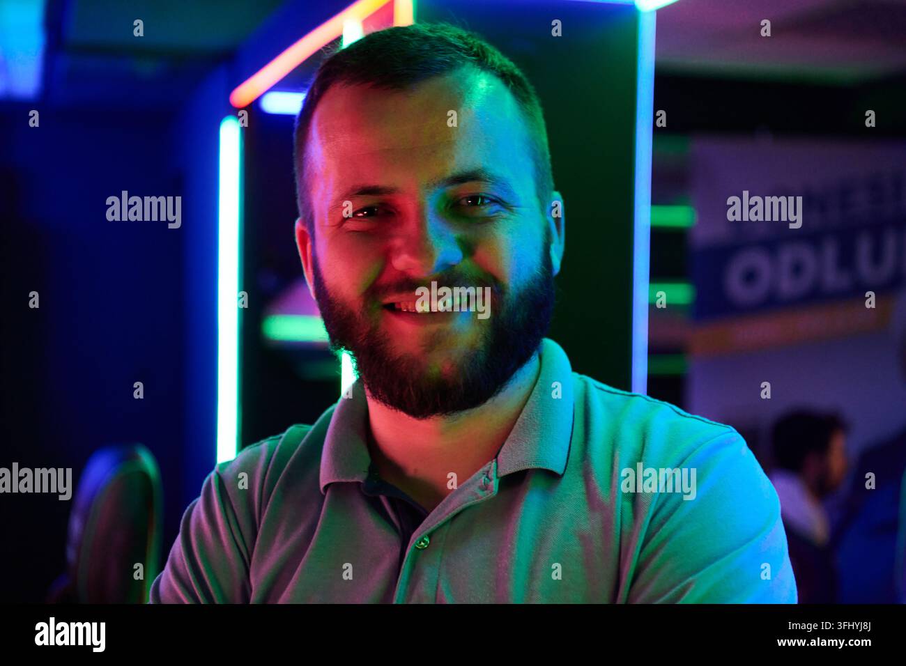Lächelnder bärtiger Mann, fotografiert in farbenfroher Neonbeleuchtung, der entspannte Selbstsicherheit und freundliche Wärme in einem lässigen Nachtleben oder Gaming-Lounge ausstrahlt Stockfoto