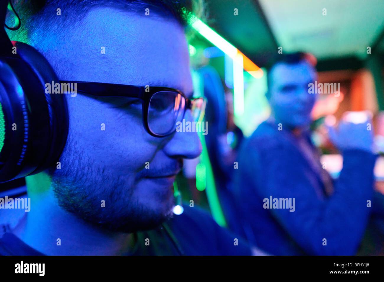 Zwei Freunde, die Seite an Seite in einem neonbeleuchteten Esports-Setup spielen, konzentriert auf das Spiel mit Headsets und Monitoren. Lebendige blaue und grüne Beleuchtung schafft ein en Stockfoto